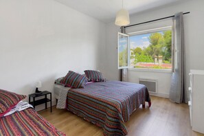 Ein Schlafzimmer mit zwei Betten, einem Fenster mit Blick auf Grünflächen, einem Nachttisch mit Lampe und einer Heizung.