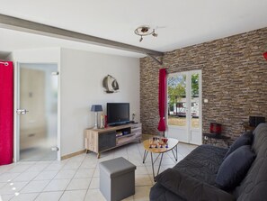A living room with a stone wall, a television, a sofa, and a door leading outside. - House and garden for 3 near the beach (La Plaine-sur-Mer)