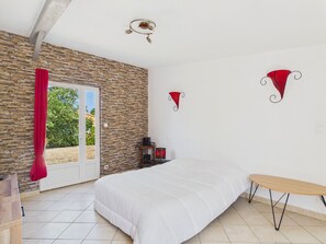 A bedroom with a bed, a bench, a stone wall, a door, and a small table with a red object on it. - House and garden for 3 near the beach (La Plaine-sur-Mer)