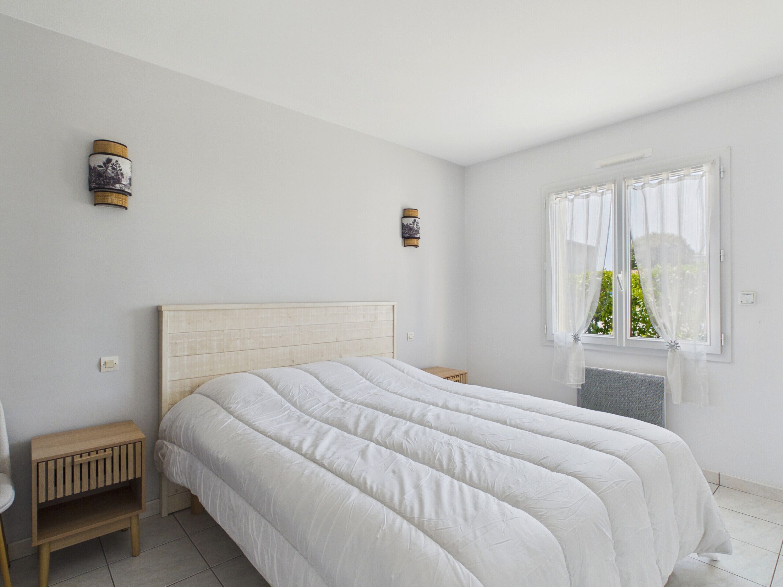 A bedroom with a large bed, a nightstand, a small wooden table, and a window with sheer curtains.