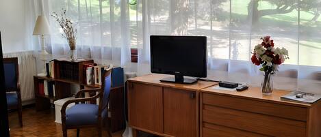 Ein Zimmer mit Schreibtisch, Stuhl, Fernseher und Blick auf einen Park durch leichte Vorhänge.