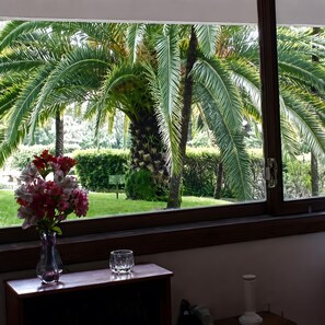 Uma janela com vista para um jardim, um vaso com flores e um copo sobre uma mesa de madeira.