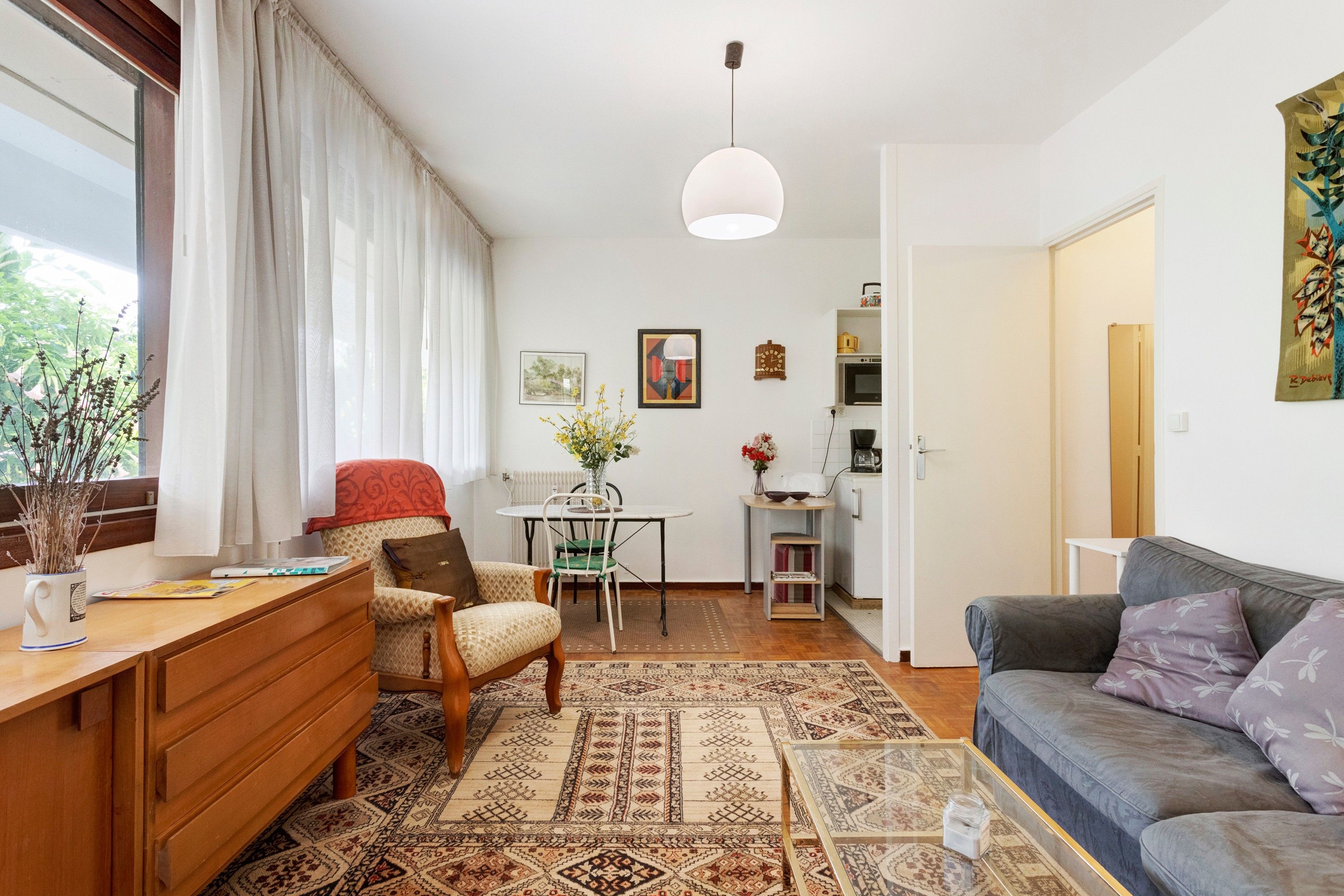 A well-lit living room with a sofa, a dining table with chairs, a kitchen area, and a large window with curtains.