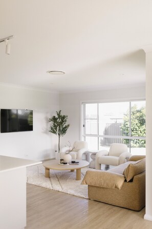 A modern living room with a flat-screen TV mounted on the wall, a round coffee table, a beige sofa, and a white armchair.
