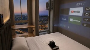 A hotel room with a bed, a bedside table, a remote control, and a large window offering a city view. - Casa Velo Residence (Teluknaga)