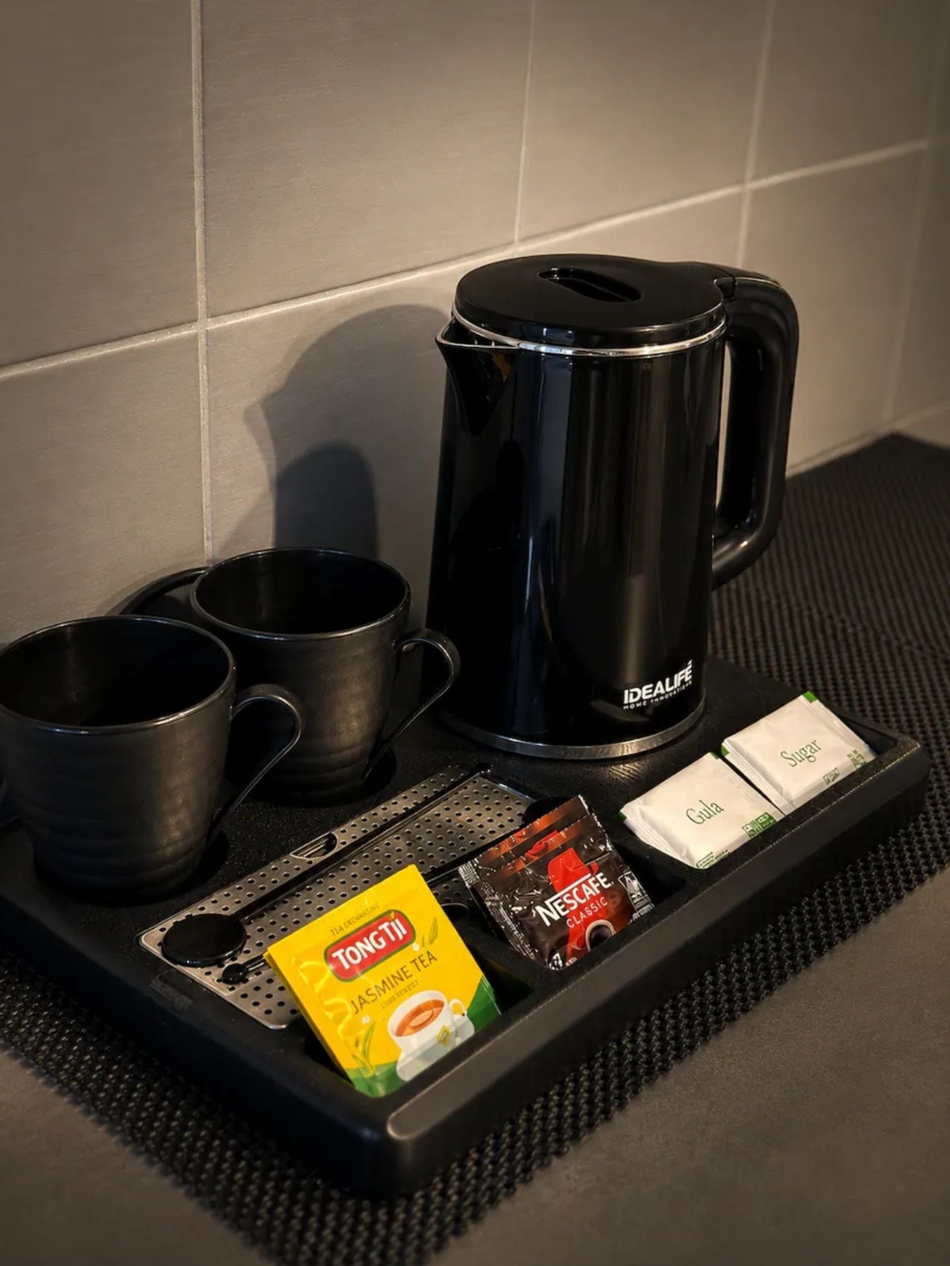 A coffee station with a black coffee maker, two black mugs, a tray with tea bags, sugar packets, and a packet of Tongari Jasmine Tea.