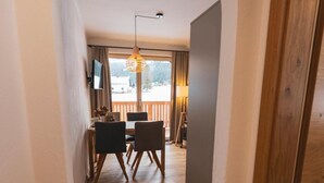 En gang med trægulv, et spiseområde med stole og et bord, samt et stort vindue med gardiner.