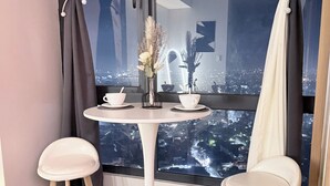 Quarto de canto com uma mesa redonda e dois banquinhos, um vaso com flores e vista para a cidade pela janela.