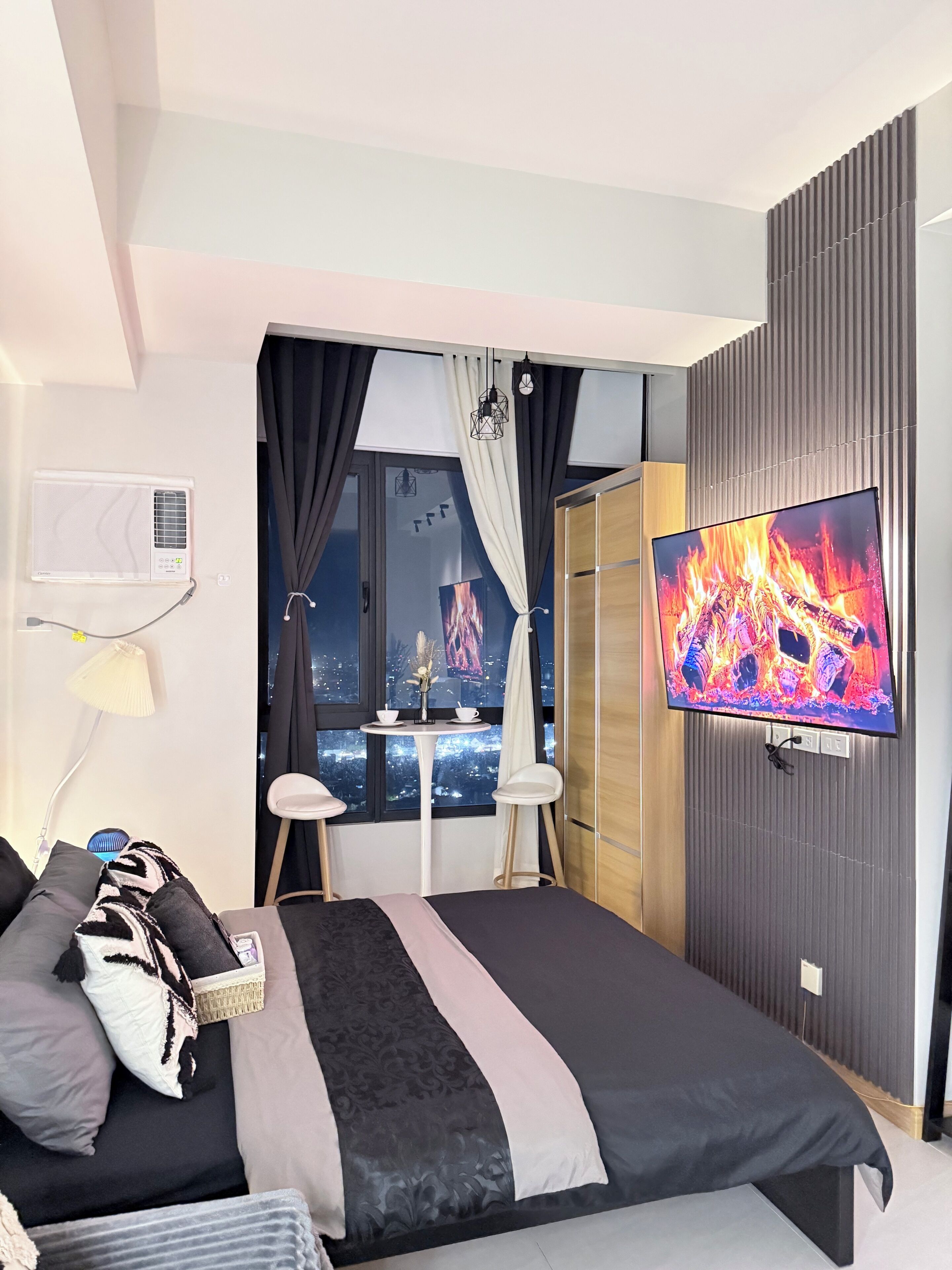 A modern hotel room with a bed, a TV, a window with curtains, a small table with a vase, and a city view.