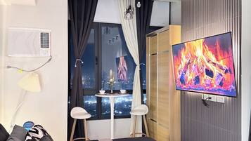 A modern hotel room with a bed, a TV, a window with curtains, a small table with a vase, and a city view.