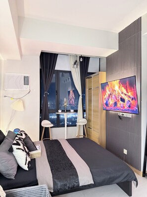A modern hotel room with a bed, a TV, a window with curtains, a small table with a vase, and a city view.