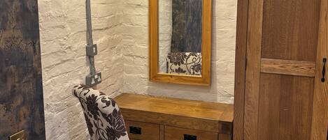 A room with a wooden cabinet, a chair with a patterned cushion, a bedside table with a lamp, and a wall-mounted mirror.