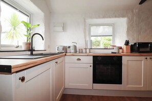 A kitchen with white cabinets, a wooden countertop, a sink, a toaster, a kettle, a microwave, and a window with blinds. - Primrose Cottage (Redruth)