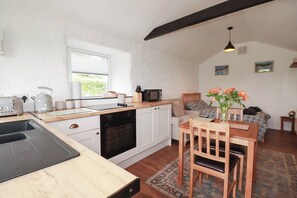 A compact kitchen with a dishwasher, a stove, and a microwave. There is a dining area with a table and chairs, and a small rug on the floor. - Primrose Cottage (Redruth)