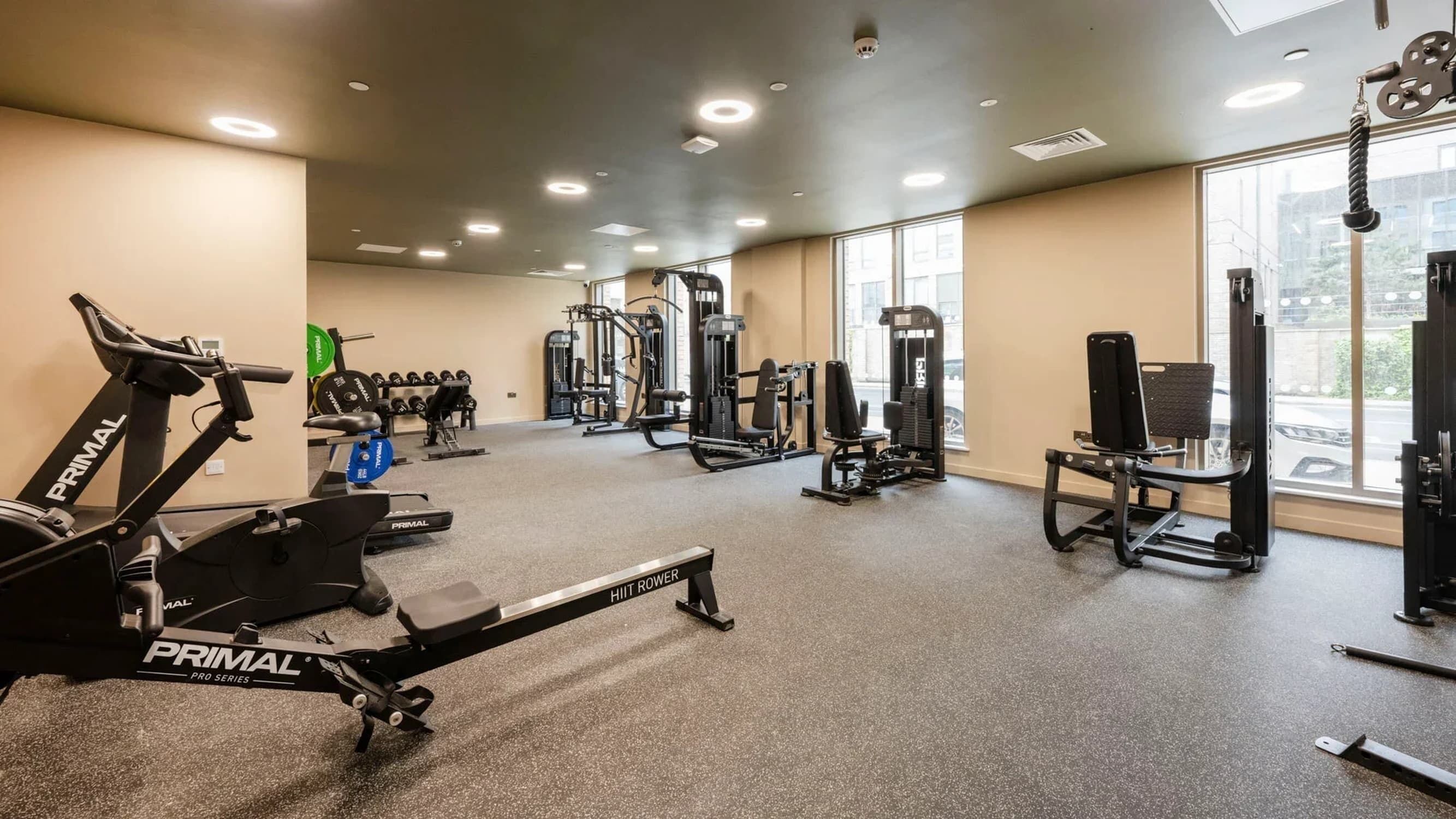 Un gimnasio con diversos equipos de ejercicio, incluyendo cintas de correr y máquinas de pesas, y amplias ventanas que ofrecen luz natural.