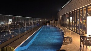 Ein Pool auf dem Dach mit Glasgeländer, ein Essbereich mit Tischen und Stühlen sowie ein Blick auf die nächtliche Skyline.