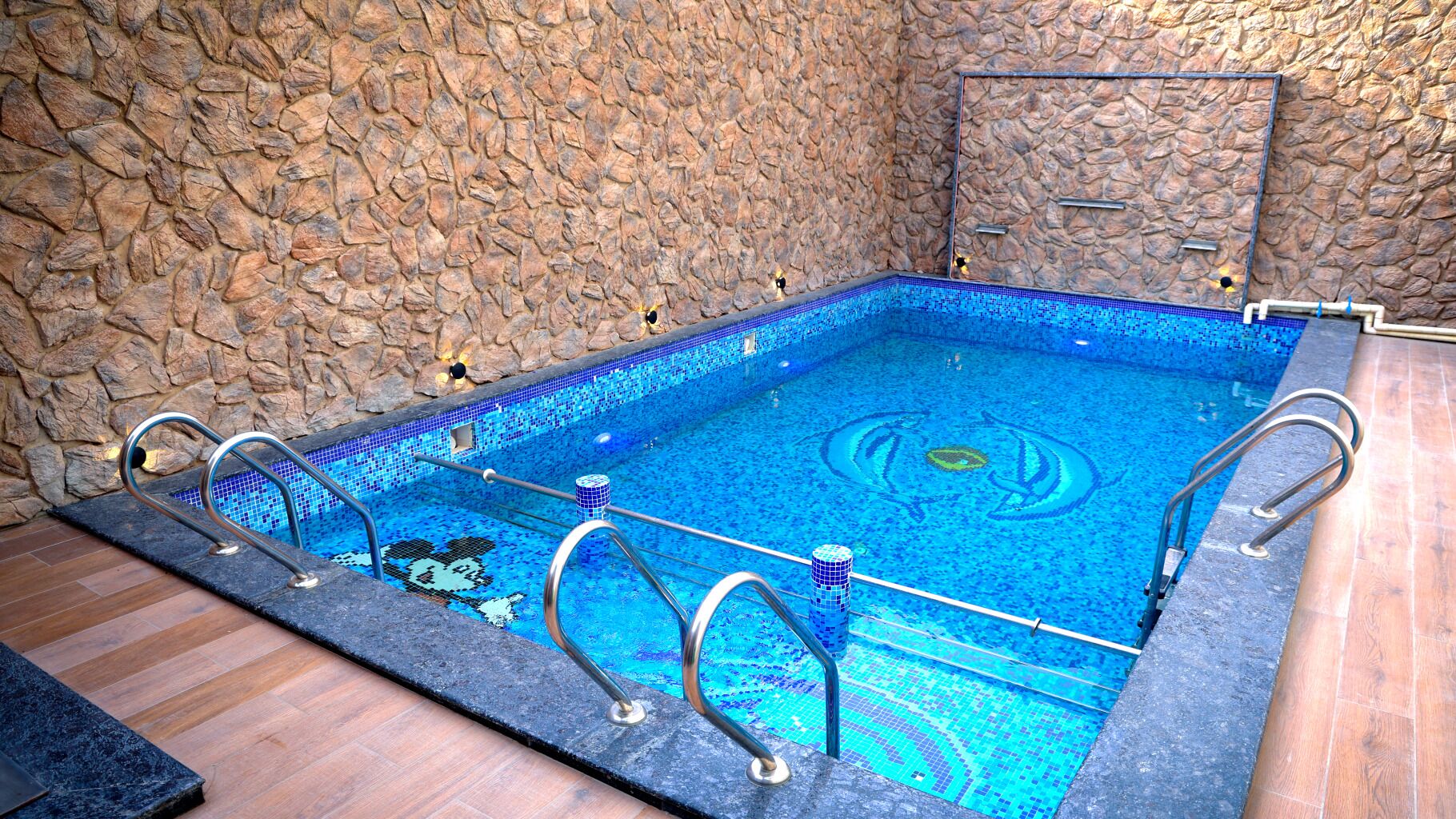 Piscina rectangular con diseño de mosaicos, rodeada de muros de piedra y piso de madera.