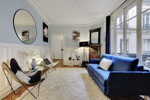 Ein Wohnzimmer mit einem blauen Sofa, einem runden Spiegel, einem Esstisch mit Stühlen und einem großen Fenster mit Blick auf die Stra ße.