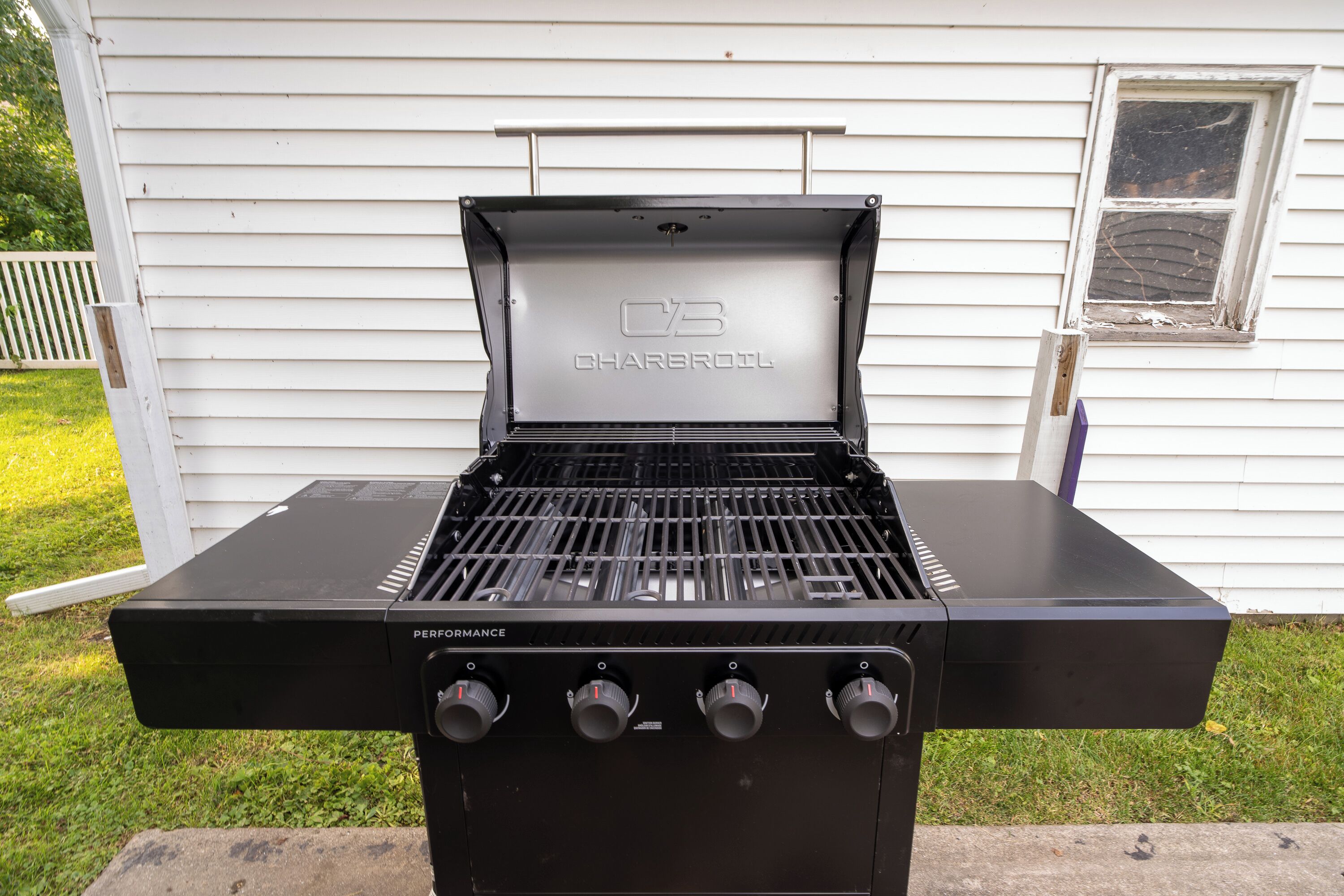 A charcoal grill with four control knobs, branded 'CHARBROIL,' positioned in front of a white house with a window.