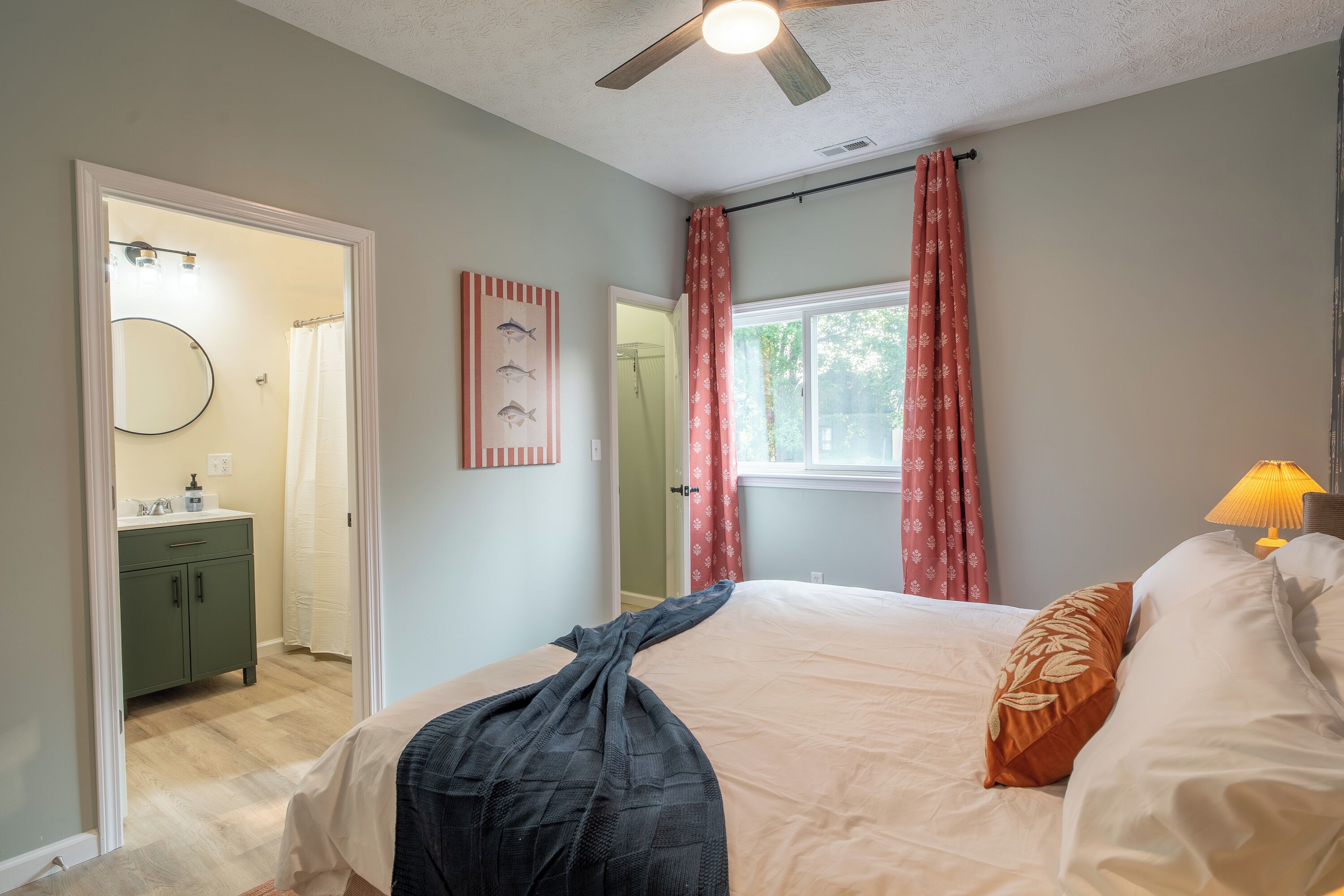 A bedroom with a bed, a ceiling fan, a window with curtains, a bedside lamp, and a bathroom with a mirror and sink.