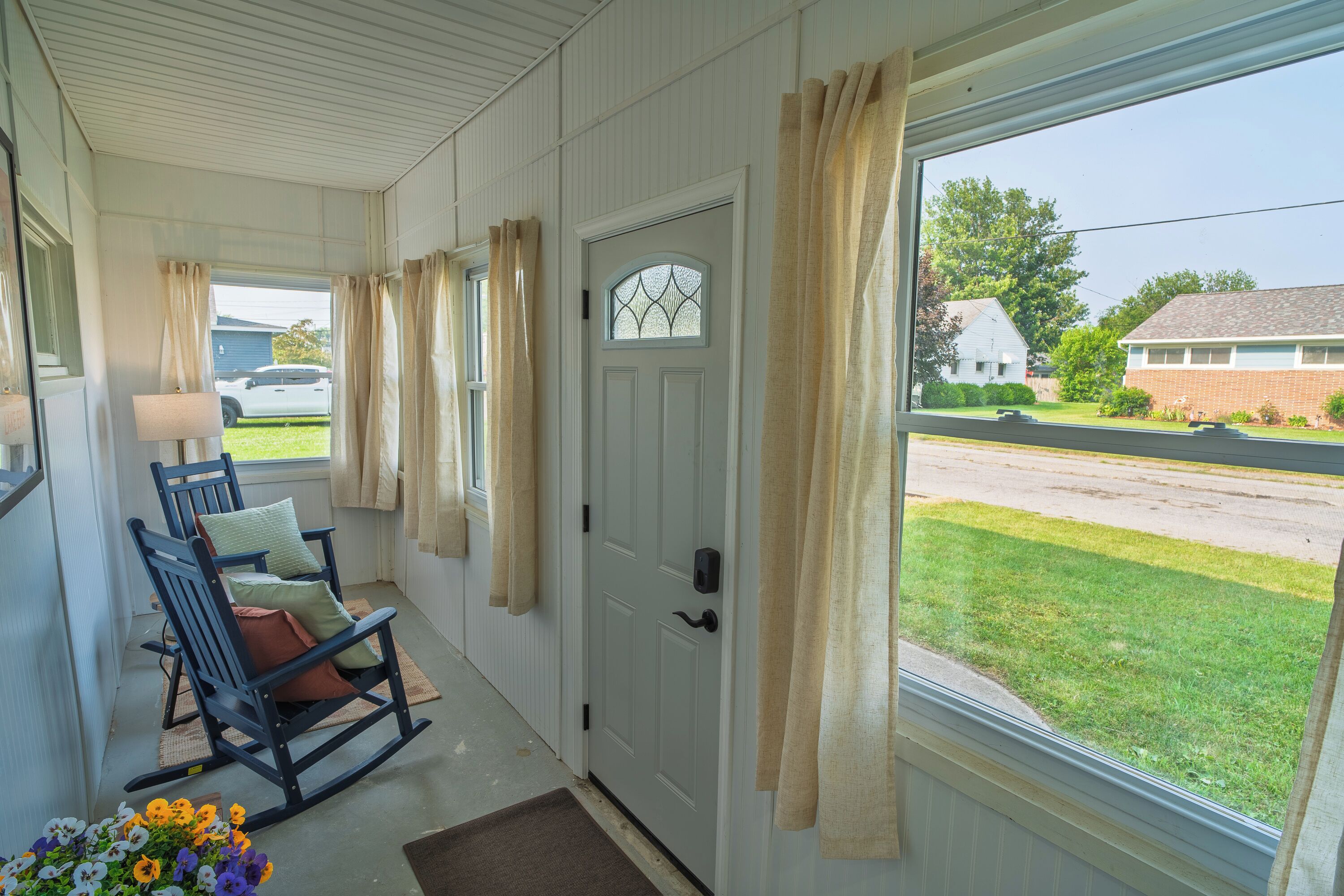 A porch with a rocking chair, a door with a window, and a view of a street and houses.