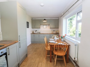 A kitchen with a dining table set for four, wooden chairs, and a window with a view of greenery. - Wilpol (Llanerchymedd)
