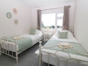 A bedroom with two single beds, a small bedside table, a window with curtains, and wall decorations. - Wilpol (Llanerchymedd)