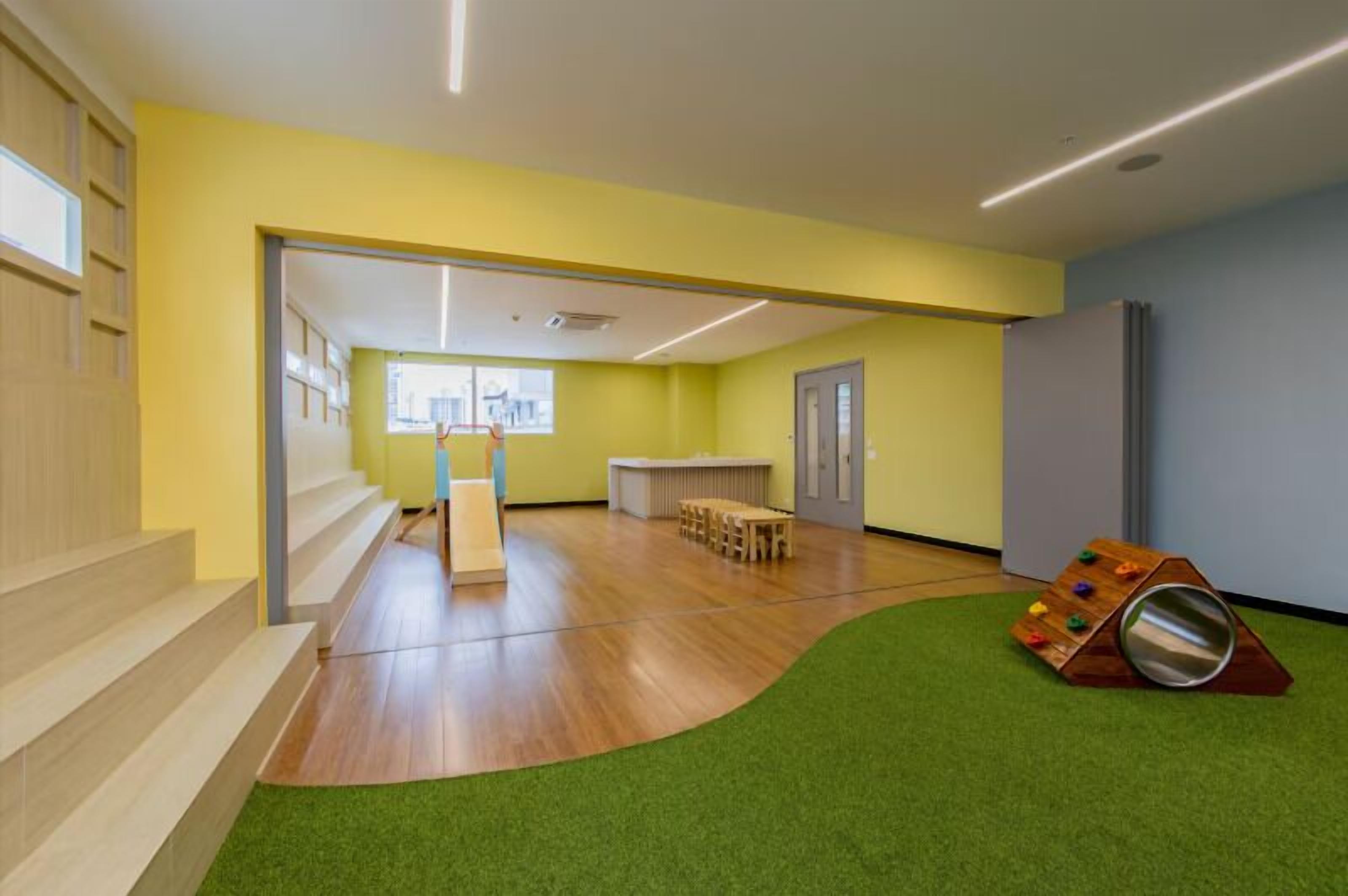 A room with a wooden floor, a green carpet, a play area with a slide, and a window providing natural light.