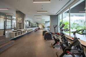 Une salle de sport équipée de vélos d’appartement, de bancs et d’une grande fenêtre donnant sur un espace verdoyant.