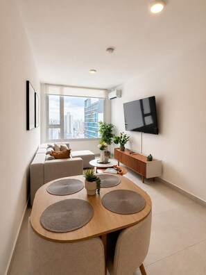 A modern living room with a sofa, a dining table with chairs, a TV mounted on the wall, and a window with a city view. - Charming 2BR + 2BA in Panama City's most luxurious building ✨🏙️🏊 (El Cangrejo)