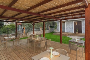 Un patio al aire libre con mesas y sillas de madera, un muro de piedra y vistas a una casa.