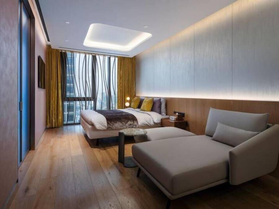 A modern hotel room with a bed, a sofa, a nightstand, and a large window with curtains.