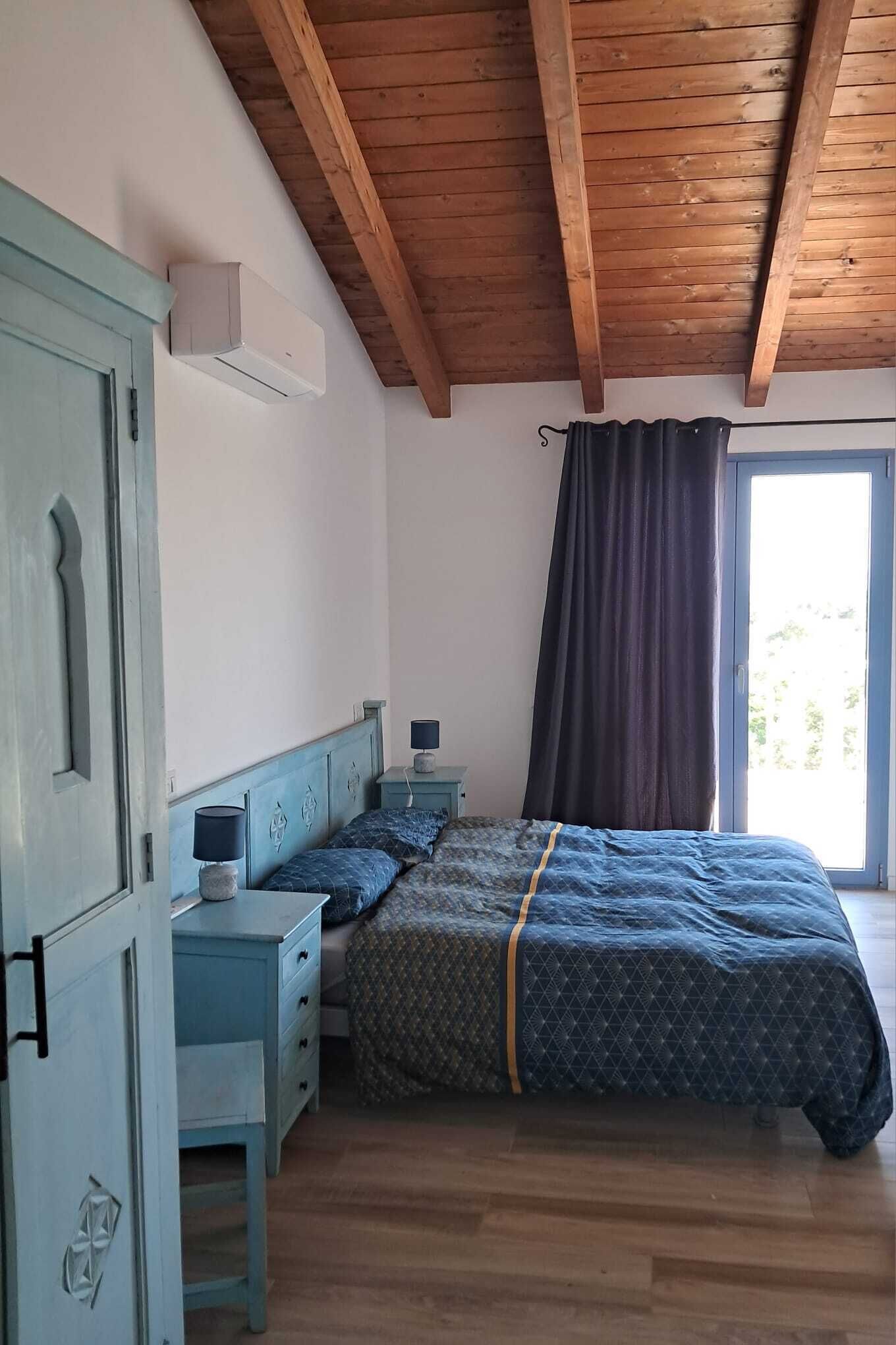 Une chambre avec un plafond en bois, un lit recouvert d’une couvre-lit à motifs, une table de chevet, une commode et une fenêtre avec des rideaux.