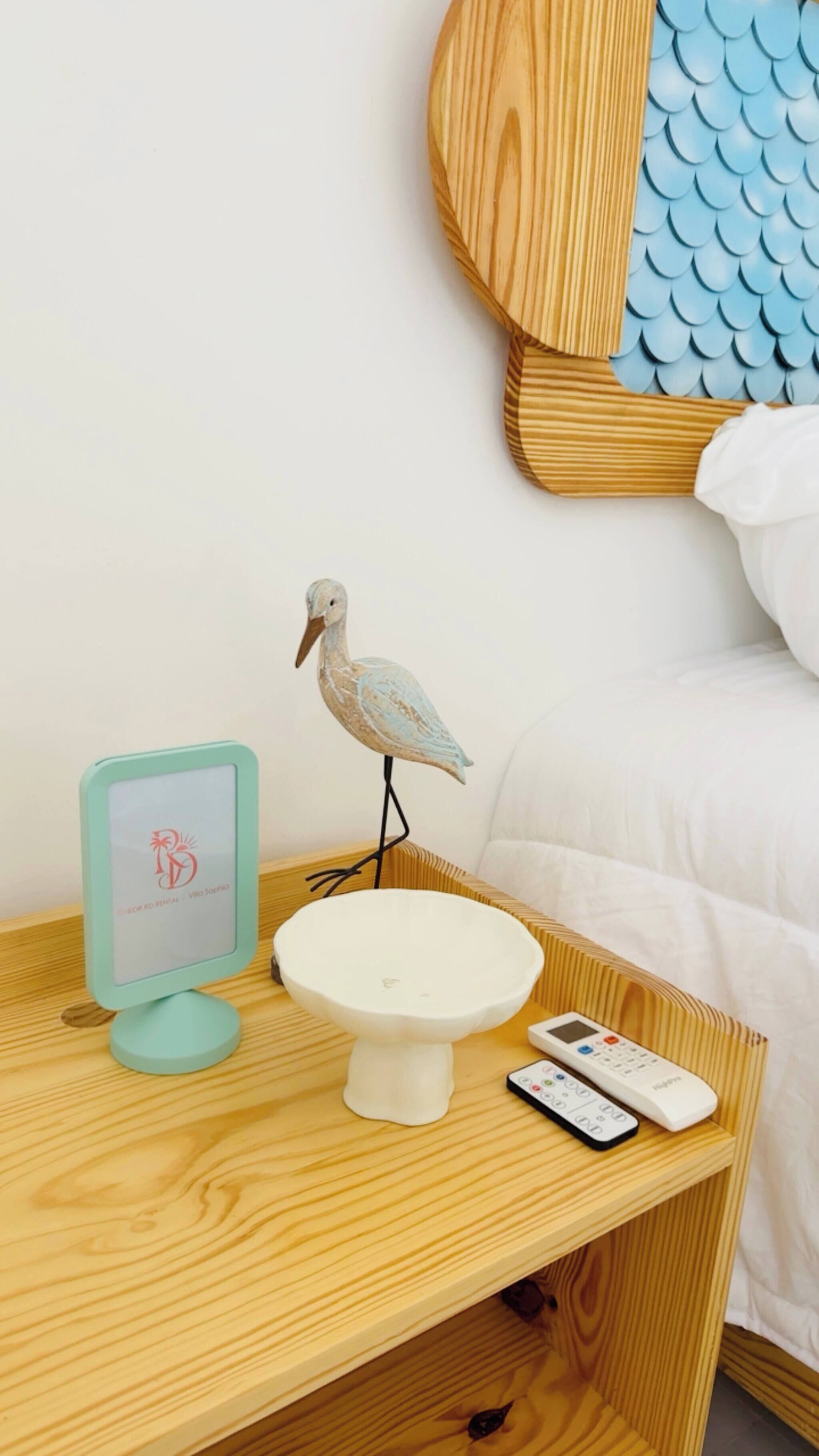 Una mesita de noche de madera con un pájaro decorativo, un cuenco blanco pequeño, un control remoto y un marco con una foto.