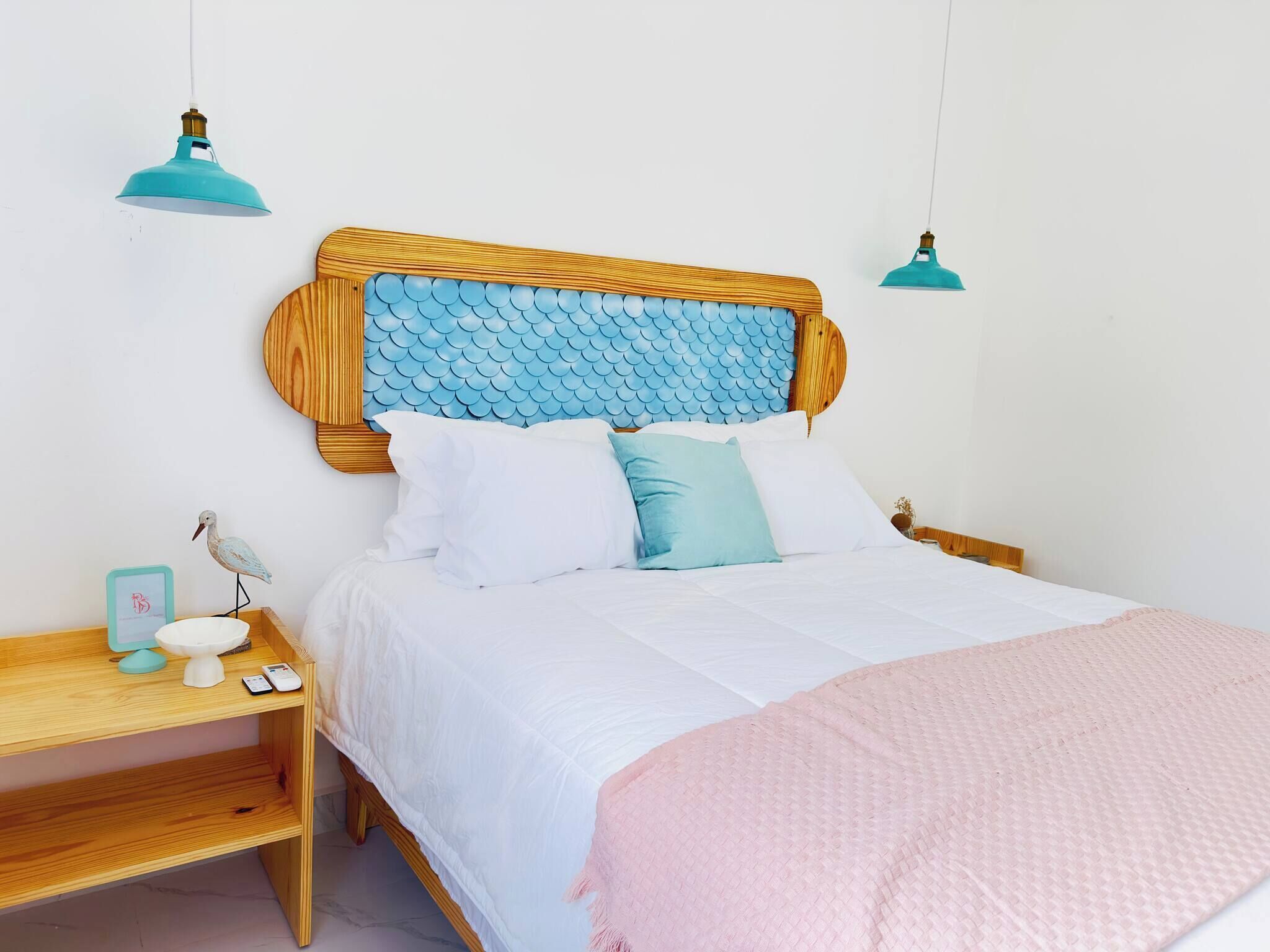 A bed with a wooden headboard, white bedding, and a pink throw blanket. There is a bedside table with a lamp and a small decorative item.