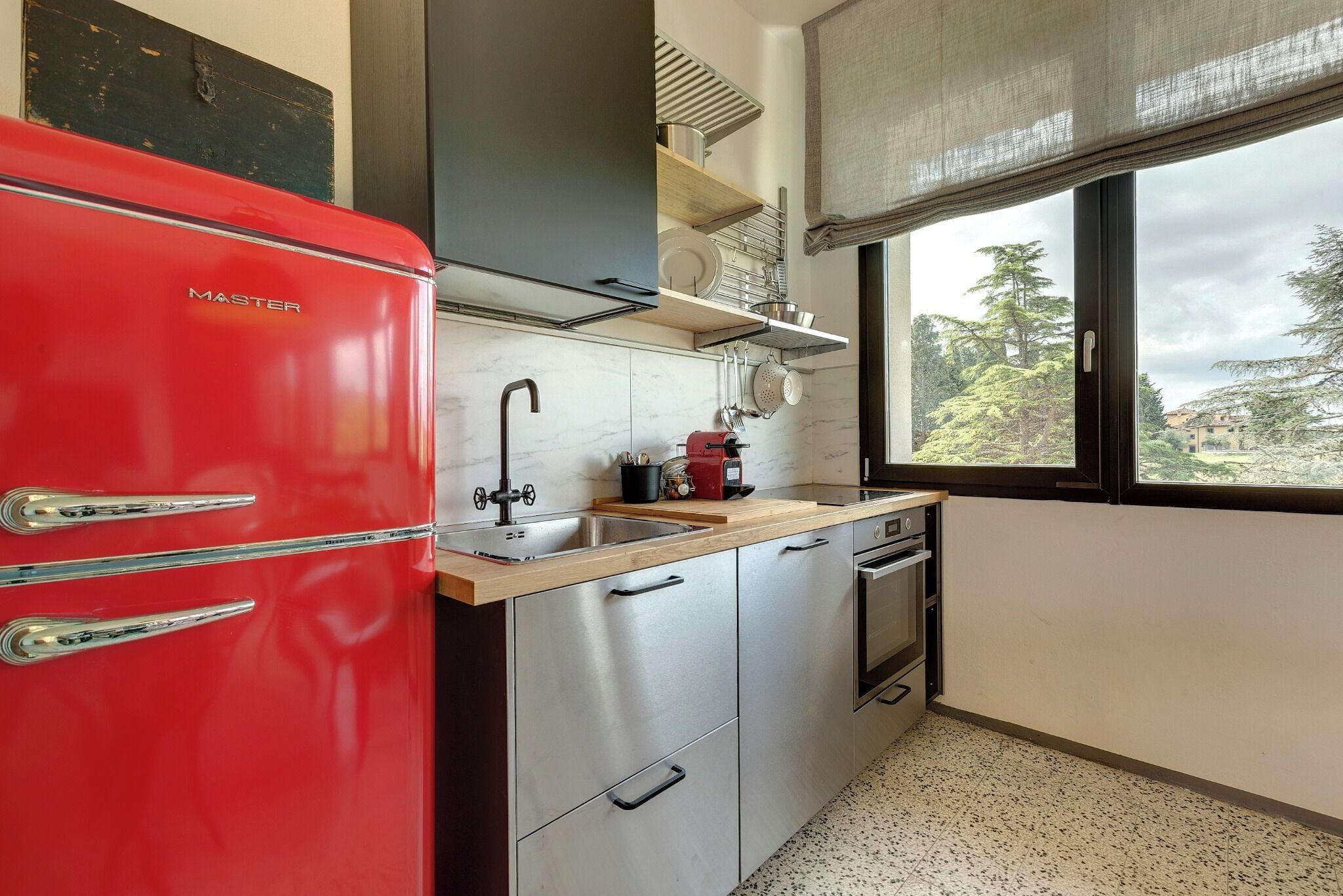 Une cuisine compacte avec un réfrigérateur rouge, des appareils en acier inoxydable et une fenêtre donnant sur des arbres et des immeubles.