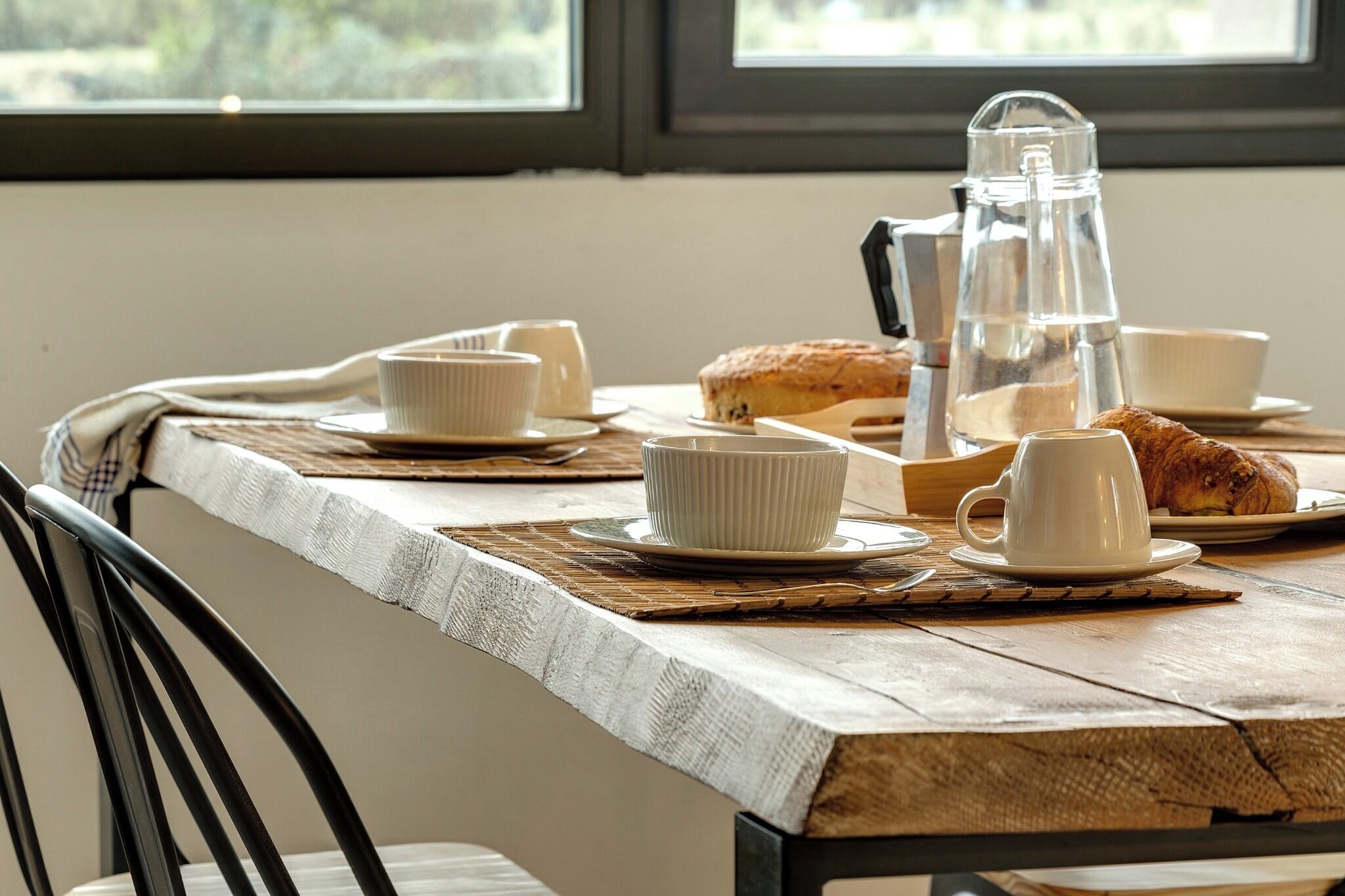 Une table à manger dressée avec de la vaisselle en céramique blanche, une carafe en verre et un croissant.