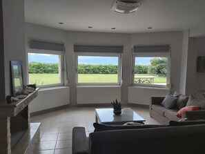 A living room with a sofa, a coffee table, and a view of a green landscape through large windows. - Villa Marilena at Nafpaktos. (Paleopanagia)