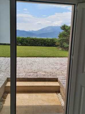 A view through a glass door leading to a paved outdoor area with a stone pathway, a grassy lawn, and a mountain backdrop. - Villa Marilena at Nafpaktos. (Paleopanagia)