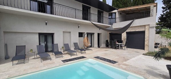 A modern outdoor pool area with lounge chairs, a dining table, and a hanging chair. - Family Home in Provence (Piolenc)