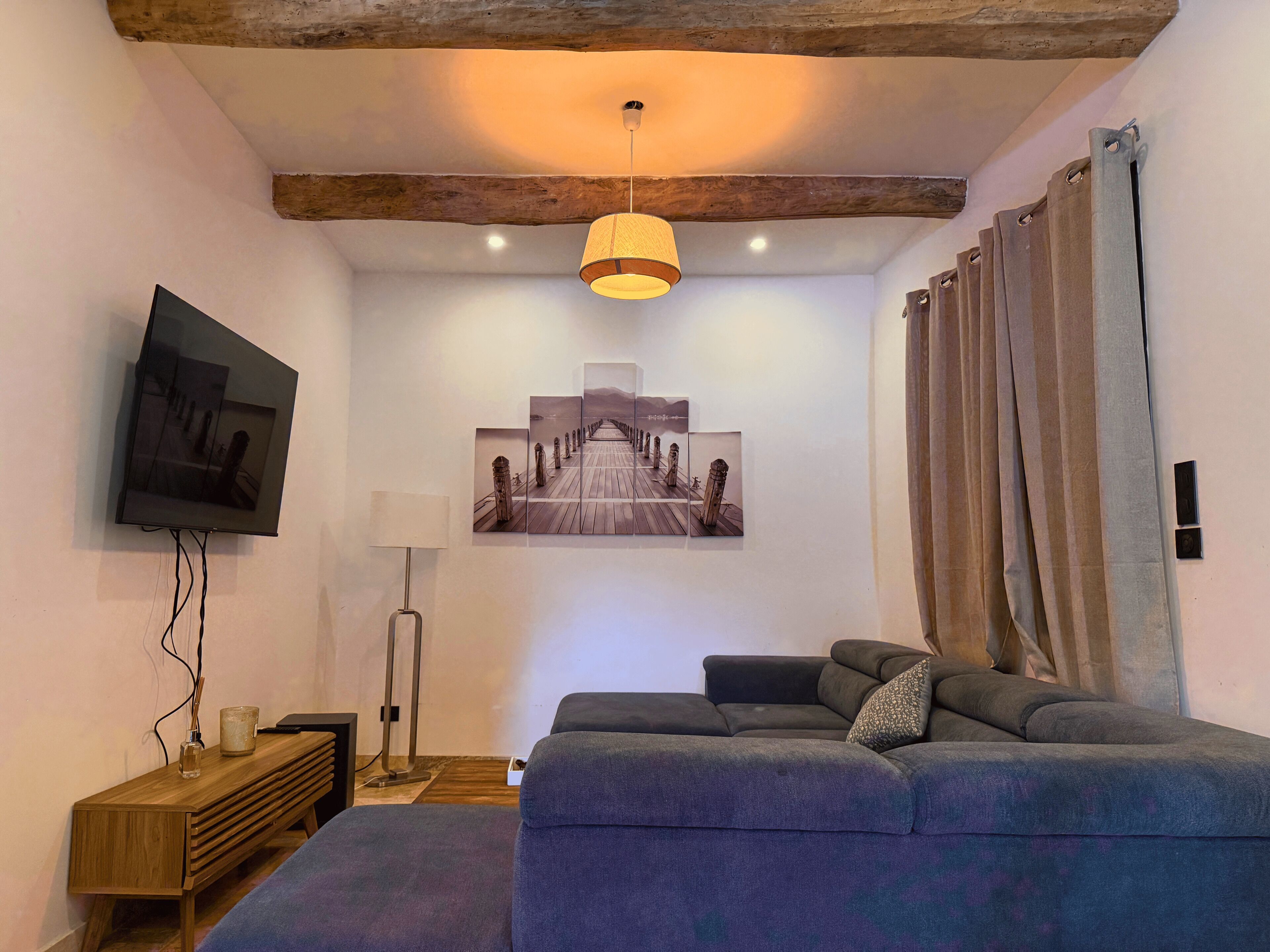 A modern living room with a sofa, a flat-screen TV, a painting, and a wooden ceiling beam.