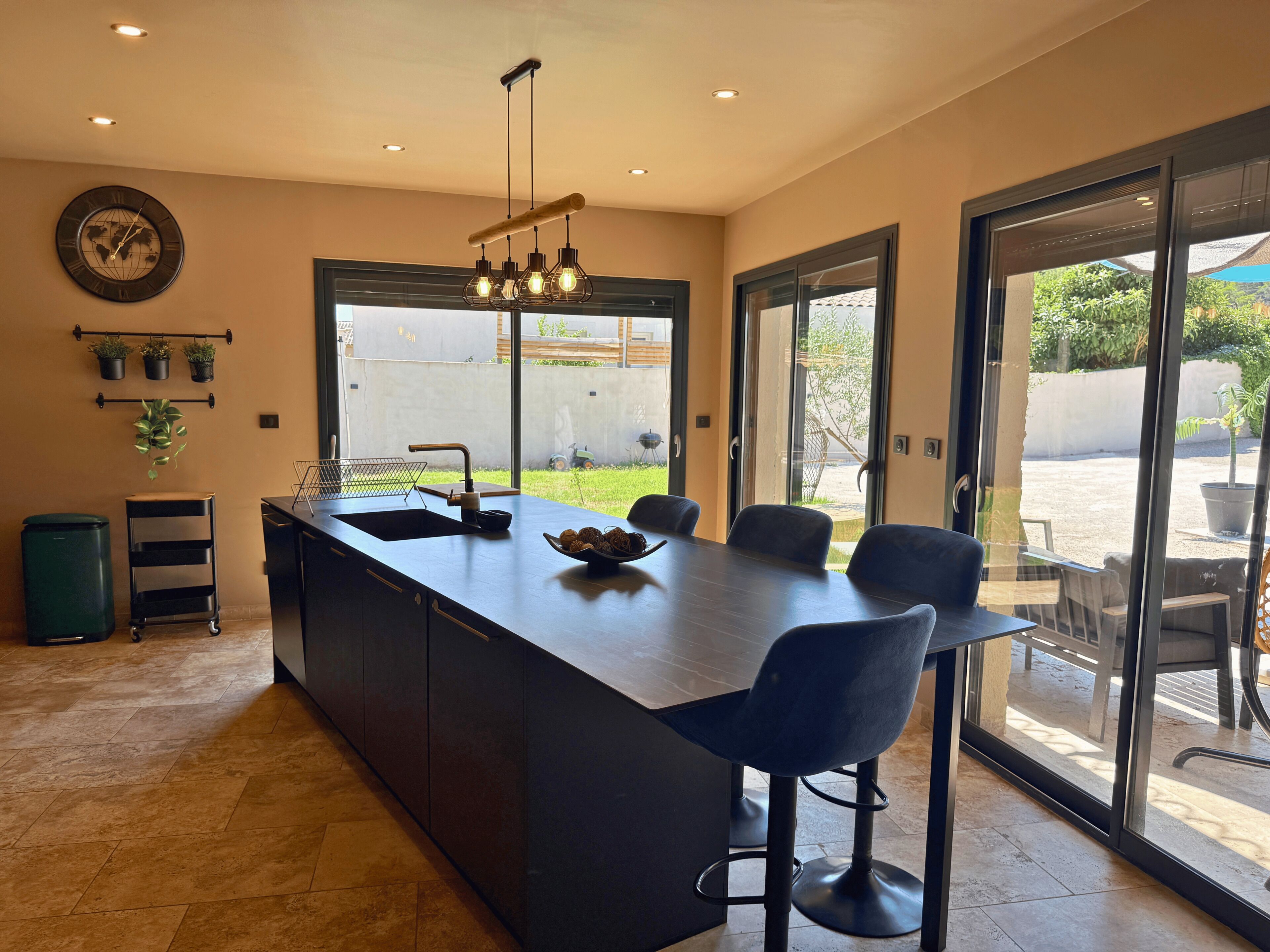 A modern kitchen with a central island, bar stools, a large window, and a view of the outdoors.