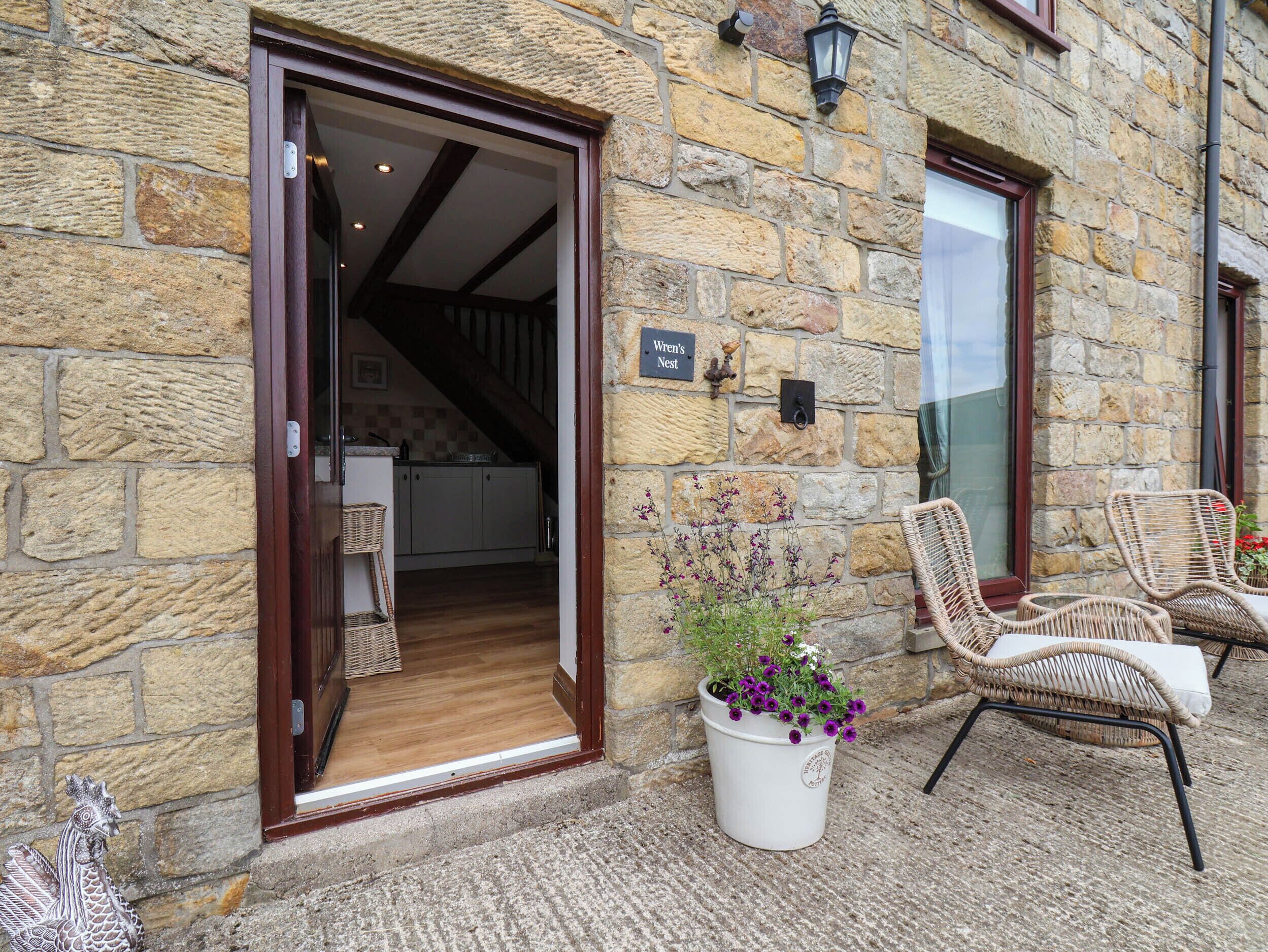 A stone building with a wooden door leading to a kitchen, a sign reading 'Wren's Nest,' and wicker chairs outside.
