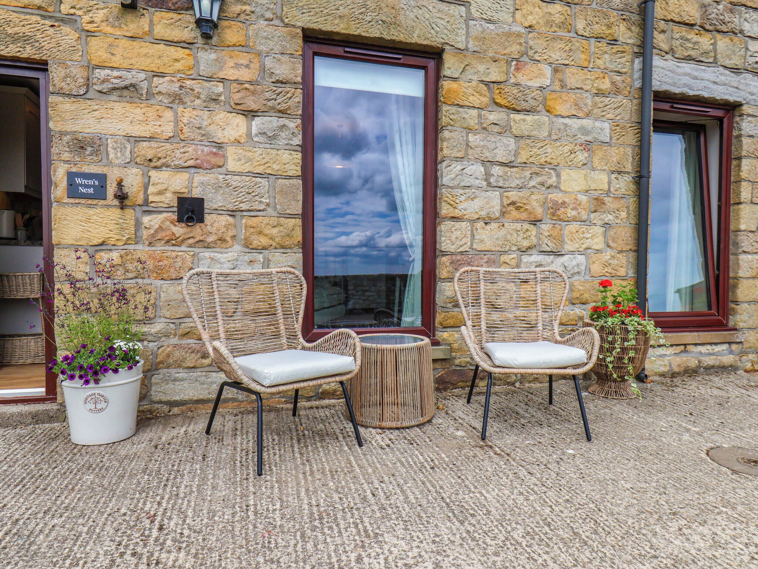 A stone building with two wicker chairs and a small table, a potted plant with purple flowers, and a sign reading 'Wren's Nest.'