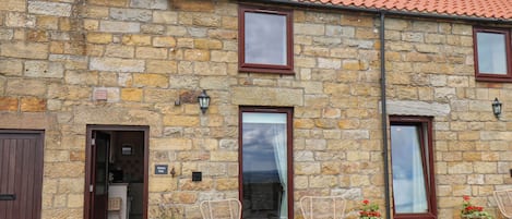 A stone building with a tiled roof, a wooden door, and a window with a curtain.