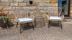 Two wicker chairs with cushions, a small round table, and a stone wall with a window reflection. - Swallow Cottage (Whitby)