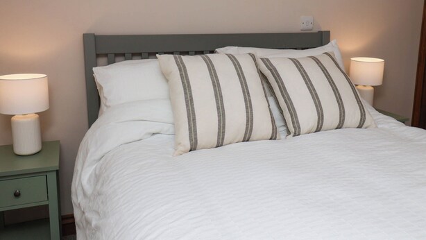 A neatly made bed with white bedding and striped pillows, flanked by two bedside tables with lamps. - Swallow Cottage (Whitby)