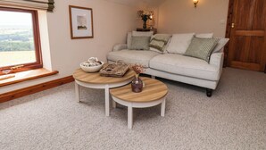 A living room with a sofa, two round tables, a window with a view, a door, and a wall-mounted light fixture. - Swallow Cottage (Whitby)
