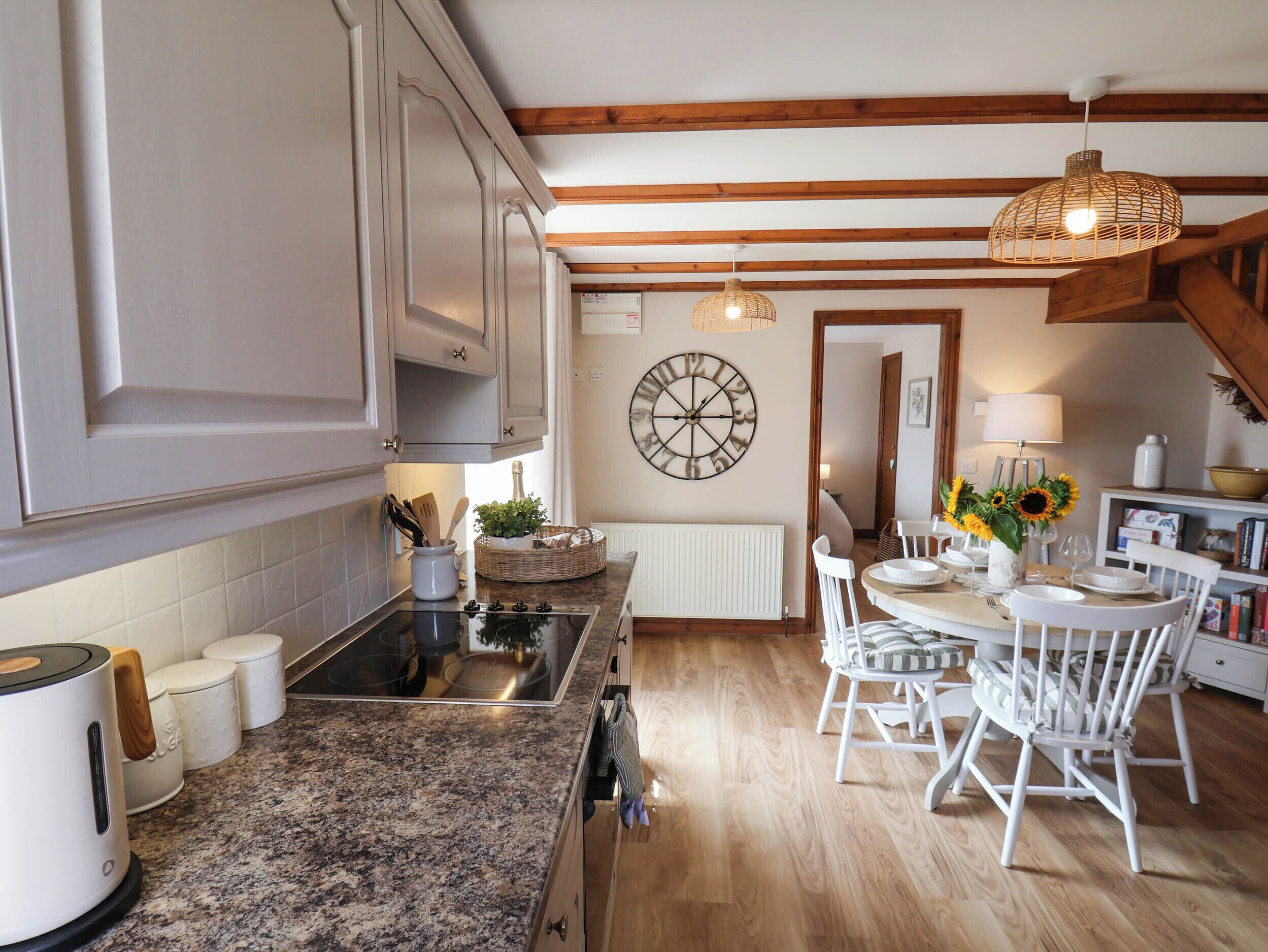 Una cocina con una isla central, armarios blancos, un reloj de pared redondo y un comedor con sillas blancas y una mesa dispuesta para dos.