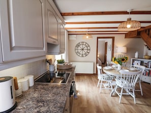 A kitchen with a central island, white cabinetry, a round wall clock, and a dining area with white chairs and a table set for two. - Swallow Cottage (Whitby)
